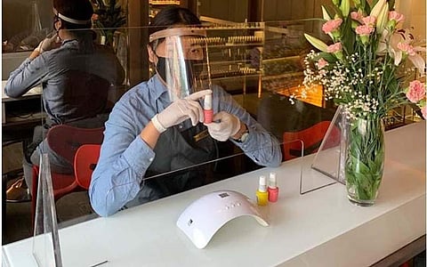 A manicurist wear protective gear and sits behind a sneeze-proof divider at Blown